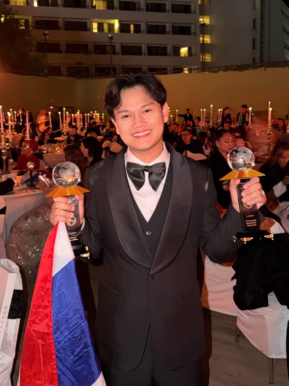 อาจารย์ทีนถือถ้วยรางวัล 2 ใบ พร้อมธงชาติไทย ในงานประกาศรางวัล